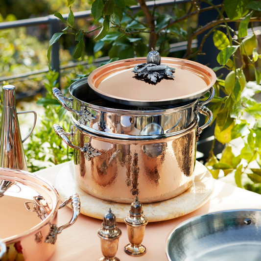 A Ruffoni hammered copper Pasta Pot with Stainless Steel Pasta Insert displayed outdoors on a table, with green foliage in the background, and matching cookware partially visible.