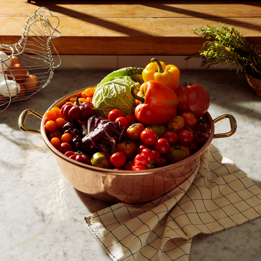 A Ruffoni Historia copper Jam Pot with side handles, filled with assorted fresh vegetables including tomatoes, peppers, and cabbage, set on a marble countertop with a checkered cloth underneath.