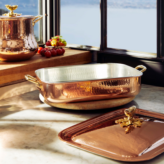 A Ruffoni hammered copper rectangular roasting pan with a tin lined interior sits on a marble countertop in a sunlit kitchen. A matching copper lid with a brass decorative handle rests in front, and other copper cookware is visible on a wooden shelf in the background near a window.
