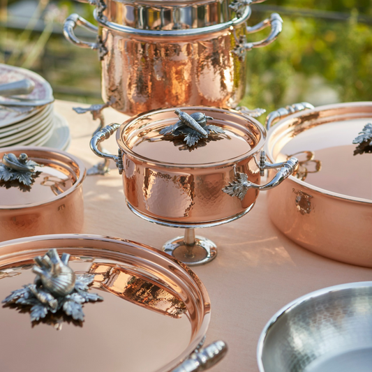 A set of Ruffoni Opus Cupra hammered copper cookware pieces with polished silver handles and knobs, arranged on a table outdoors with a blurred green garden background