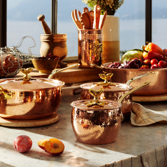 A set of Ruffoni hammered copper cookware pots and saucepans, arranged on a marble countertop in a bright kitchen setting with fruit, utensils, and a window view in the background