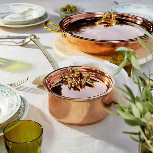 Two Olivia Cupra copper pots with brass handles, styled on a white tablecloth with green glassware, plates, and plants in a bright dining setting