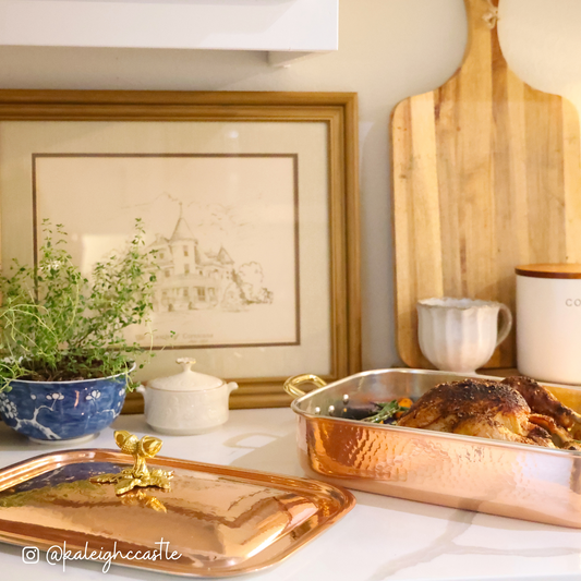 Ruffoni copper roasting pan with roasted chicken on a kitchen counter, with a framed picture and cutting board in the background photographed by @kaleighccastle