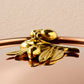 A close-up of a shiny brass decorative olive-branch knob resting on a reflective copper lid against a light beige background.