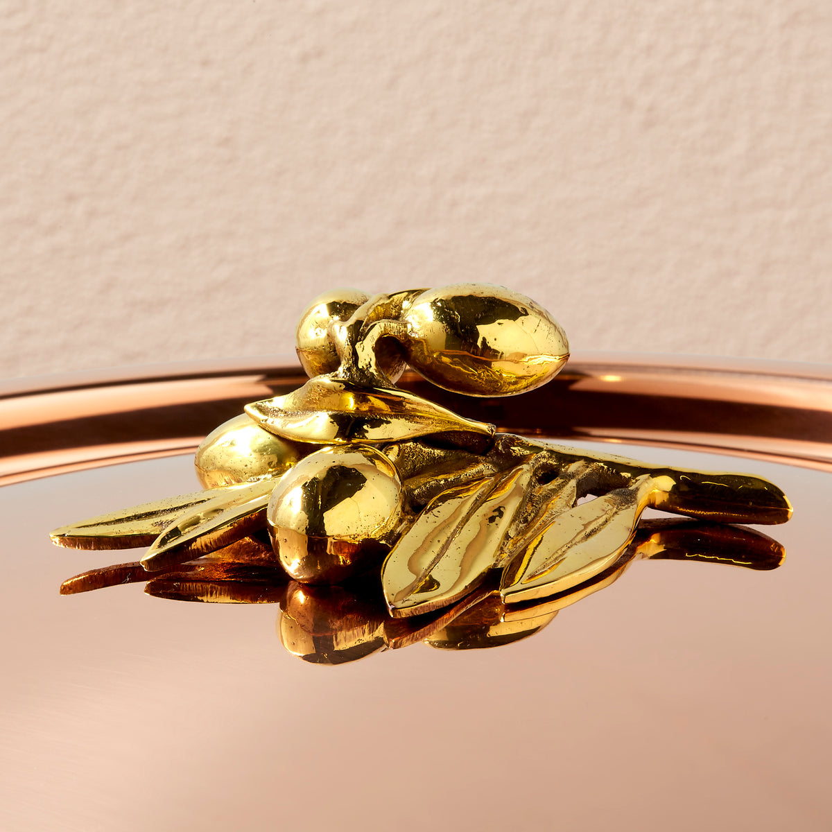 A close-up of a shiny brass decorative olive-branch knob resting on a reflective copper lid against a light beige background.