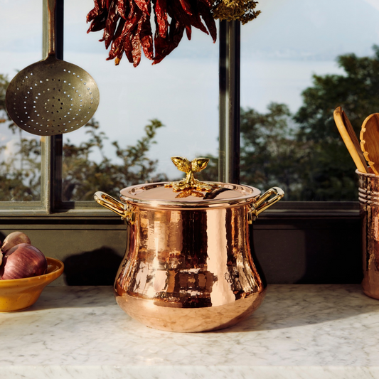 An hammered copper lidded Belly Pot with side handles sits on a marble countertop in a kitchen setting, with a window, hanging dried chilies, and other cookware visible in the background.