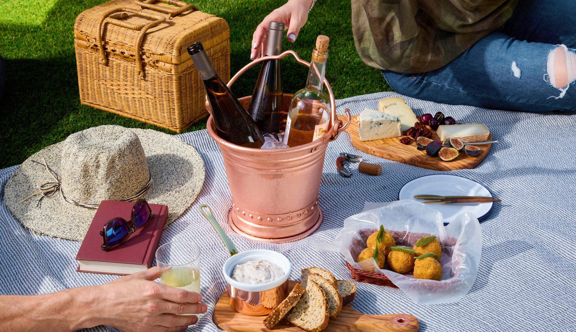 Picnic setup with wicker basket, food items, and drinks on a blanket outdoors.