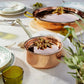Two Olivia Cupra copper pots with brass handles, styled on a white tablecloth with green glassware, plates, and plants in a bright dining setting