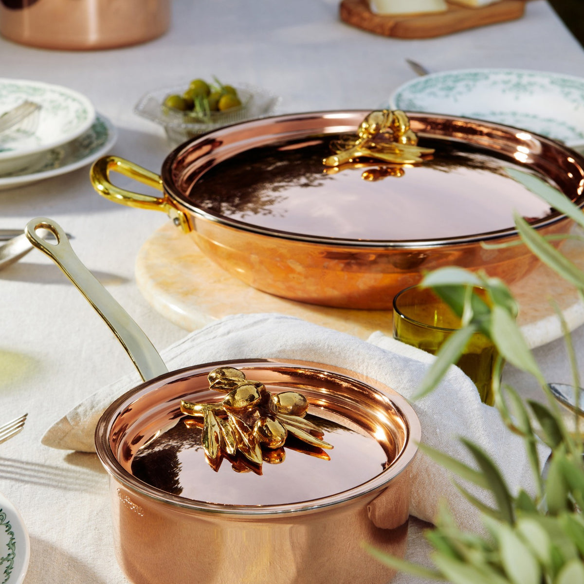 Olivia Cupra serving frying pan and saucepan on a beige linen tablecloth with green plates and glasses in the Italian countryside on a summer day
